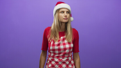 Fototapeta premium Woman wearing santa hat and red checked apron stands centered and looks to side with bare forearms visible against purple backdrop in studio; holiday boredom.