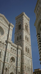 Fototapeta premium Campanile and cathedral facade standing tall with detailed green white and pink marble ornament, rose window and statues on a building in florence italy; awe.