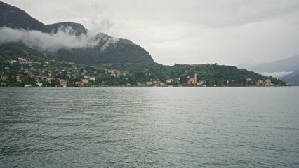Fototapeta premium Lake foreground with rippling water and shoreline village with church tower and clustered buildings at lake como, italy; serenity.