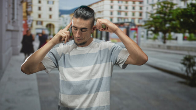 Man with manbun and striped tshirt holding smartphone to ear, gesturing with his hand while walking down a busy city street lined with buildings and trees; frustration urgency.