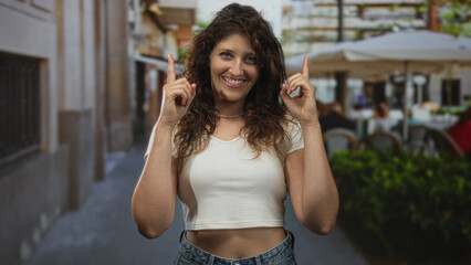 Fototapeta premium Woman points both index fingers upward on a street terrace, smiling with exposed midriff visible; joy.