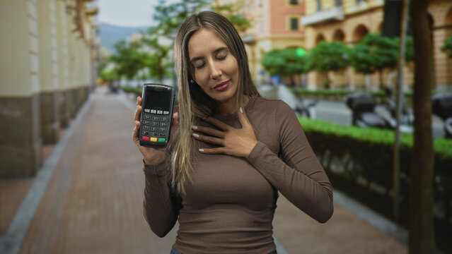 Woman young hispanic blonde holds portable card reader in city street with right hand raised showing device and left hand on chest, eyes closed; gratitude trust.