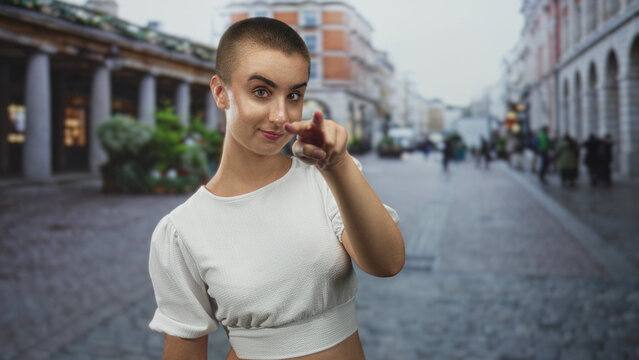 Woman with buzzcut pointing index finger forward on cobbled street in white crop top, direct eye contact and a playful scolding gesture; confident challenge.