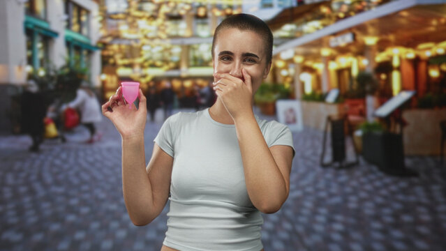 Woman with short hair holds a pink menstrual cup and covers mouth with hand on a cobblestone street; relief empowerment.
