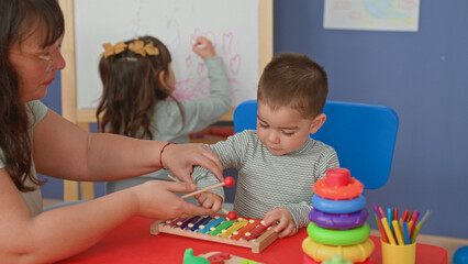 Naklejka premium Boy holding mallet tapping a rainbow xylophone on a red table with visible hand while a girl draws on an easel behind in a building; curiosity learning play.