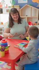 Naklejka premium Woman taps xylophone with mallet while guiding little boy at a play table in building; nurturing learning joy.