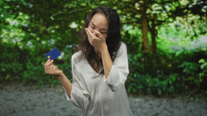 Fototapeta premium Woman holding creditcard while covering mouth with her hand in a forest, inspecting a blue card; surprise money decision.