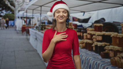 Fototapeta premium Young hispanic woman in santa hat touches hand to chest at a street market stall with jars and canopy; holiday gratitude.