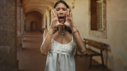 Fototapeta premium Thai woman cupping hands around mouth calling out, showing bare hands and necklace, wearing white crochet dress in a weathered building corridor; urgent appeal.