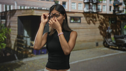 Fototapeta premium Thai woman removes eyeglasses, squints and pinches nose while standing on a street near a wooden building entrance; irritation allergy.