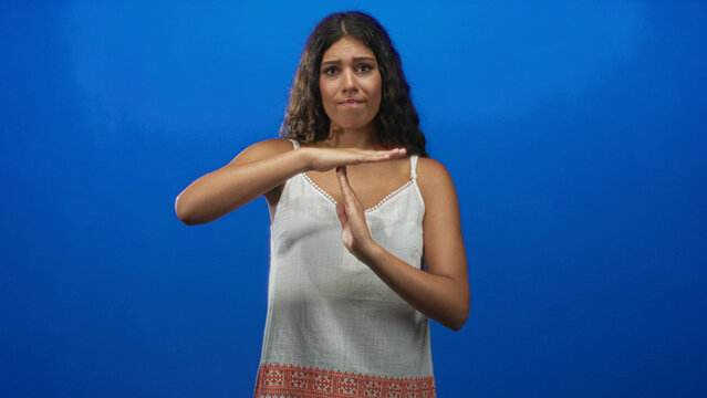 Hispanic woman in white tanktop making timeout sign with hands in studio with blue backdrop and soft lighting, embroidered hem visible; calm reflection.