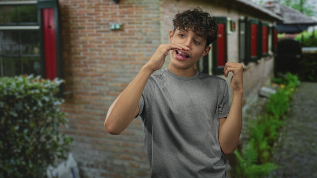 Teenage boy gesturing with hand and miming speech in front of brick building window, casual tshirt and courtyard visible; playfulness communication.