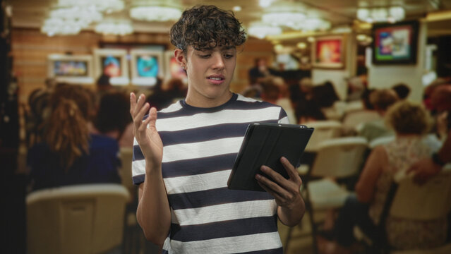 Teen boy in striped shirt holds a tablet with one hand and presses his bare hand to chest while reading aloud in a crowded building; anxiety reflection.