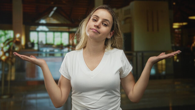 Woman in white tshirt standing palms up shrug at hotel lobby reception area, casual pose and neutral expression; indifference doubt ambivalence.