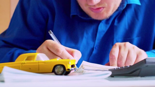 Slow zoom out on car dealer  calculating and writing data in ledger. Focus on hands, yellow toy car on desk. Close up, cinematic shallow depth of field, natural daylight.