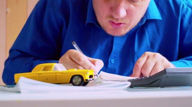 Focused car businessman using calculator and writing notes in ledger. Focus on hands.Yellow toy car on table. Shallow depth of field, natural daylight. 