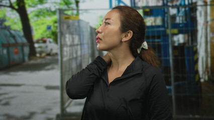 Fototapeta premium Woman with hand on neck near metal fence on street, leaning by dumpsters and trees with visible neck skin; discomfort fatigue.