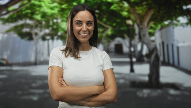 Woman with crossed arms showing hands and forearms smiling confidently on street lined with trees and building facades; confidence approachability.