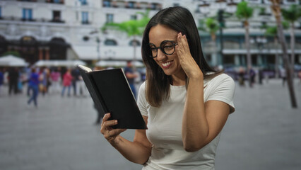 Fototapeta premium Woman reading a black book, hand raised and smiling while adjusting glasses on a busy street with people nearby; joy storytelling.