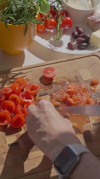 Hand removes pulp from cherry tomatoes with knife on board, with herbs, olives and garlic nearby. Concept of appetizer and bruschetta preparation