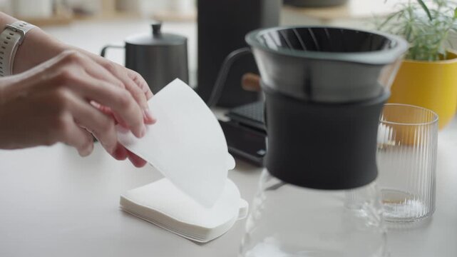 Hands preparing paper filter for pour over coffee beside dripper and glass carafe. Concept of manual coffee brewing