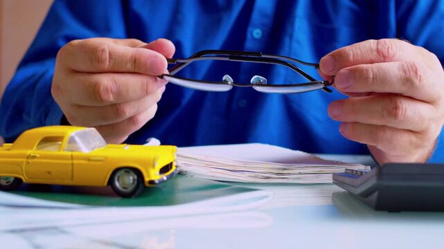 Car insurance agent  twirling glasses at office desk close up. Focus on hands and eyeglasses, shallow depth of field, natural daylight. Bored businessman procrastinating at work.