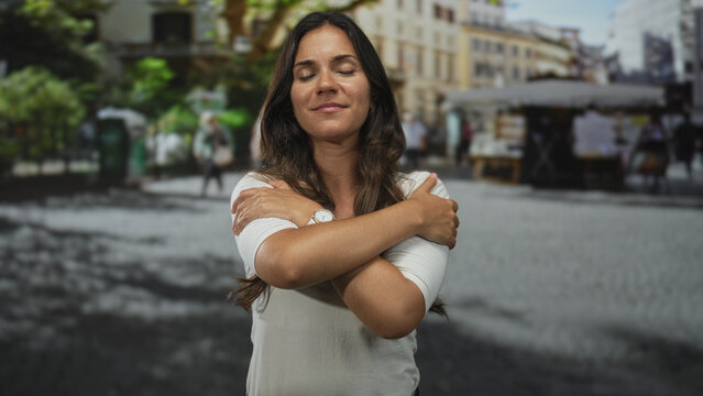 Woman crossing arms in a self hug on a city street with eyes closed and a soft smile; self care calm.