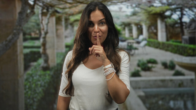 Woman with finger to lips shushing near colonnade and fountain in building garden park, white shirt and bracelets visible; secrecy quiet.