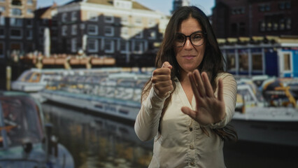 Fototapeta premium Woman wearing glasses with hands forward in stop gesture and steering motion on street near canal boats and buildings; playful travel.