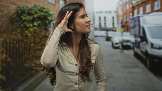 Woman cupping hand to ear on city street, leaning forward to listen for distant noise; concern curiosity.