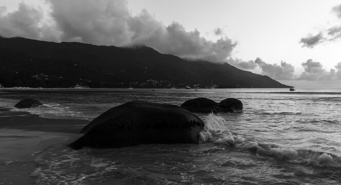 Black and white photography. Beau Vallon beach landscape taken at night. Dark shore water and silhouettes of coastal rock are under cloudy sky. Mahe island, Seychelles