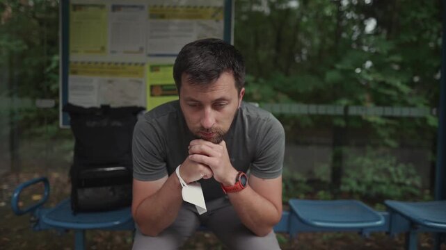 A sad, disappointed man sits at a German bus stop, checking his watch due to a delayed or cancelled public transport connection, showing frustration over transport problems and infinite waiting.