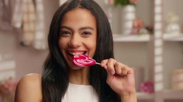 Happy smiling Hispanic ethnic girl biting sweet sugar tasty candy with white healthy teeth looking at camera Latina 20s woman American female bite lollipop smile in pink bathroom stomatology medicine