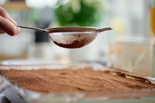 Hand holds sieve over tiramisu dessert covered with cocoa powder