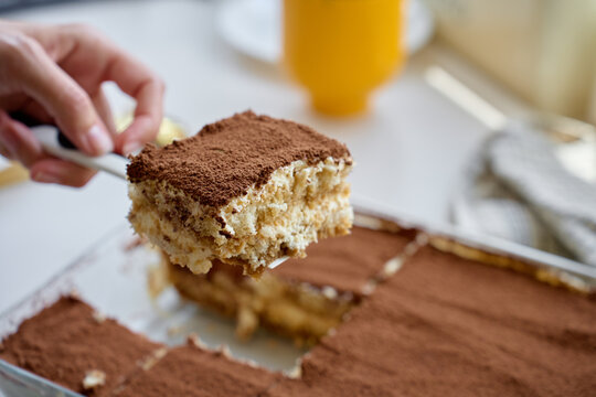 Hand lifts tiramisu slice with spatula from glass dish covered with cocoa powder