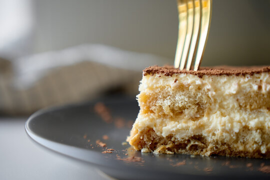 Fork presses into tiramisu slice on dark plate with cocoa topping and cream layers