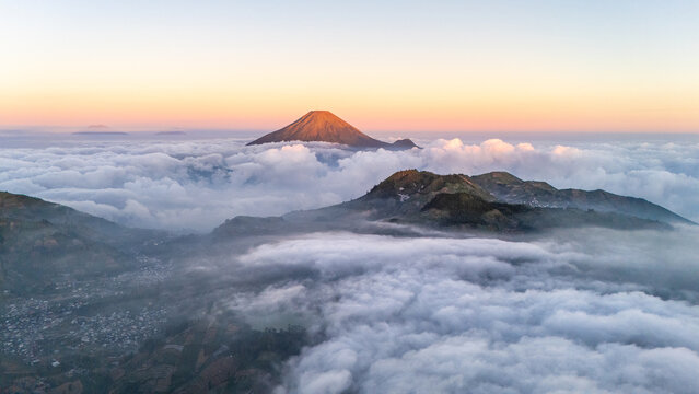 Aerial view of Mount Sindoro rising above a sea of clouds at sunrise with lush green mountain slopes and a small village visible in the valley in Indonesia.