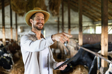 Happy farmer holding a digital tablet and pointing, managing cows and livestock inside a barn, embracing modern farm technology © Dejan