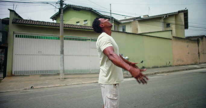 Man of African descent standing in an urban street, raising arms toward the sky, expressing gratitude and faith, symbolizing spiritual connection and hope