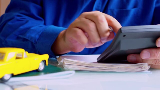 Insurance agent hands using calculator and writing in policy document close up. Yellow toy car on table, shallow depth of field, natural daylight. Calculating car coverage cost or premium.