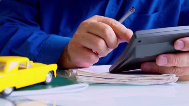 Insurance agent hands using calculator at desk close up. Focus on fingers, yellow toy car on table. Shallow depth of field, natural daylight. Calculating car coverage cost or premium.