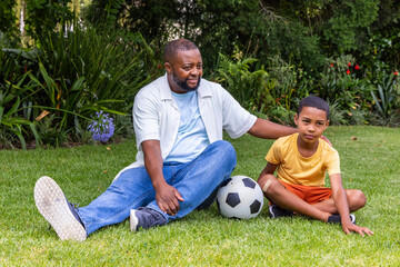 Naklejka premium African American dad and teen son sitting in garden with soccer ball, son wearing yellow shirt