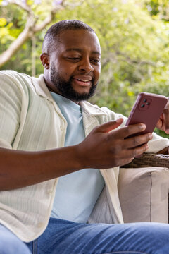 African man sitting on wicker patio chair holding maroon phone while wearing ribbed shirt and jeans