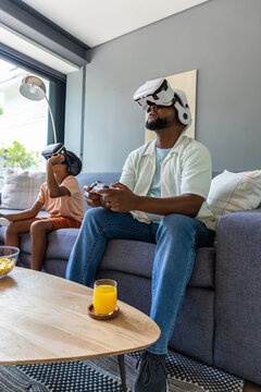 African American father and son playing VR on sofa holding gamepad and wearing white black headsets