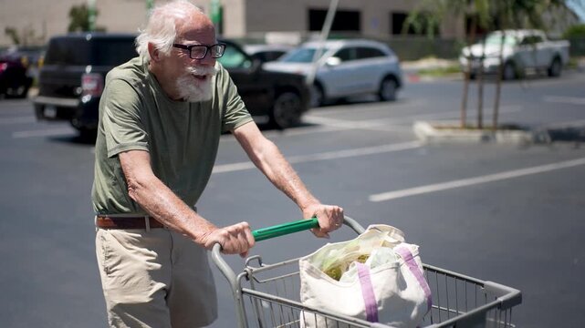 Elderly man pushes a shopping cart filled with groceries through a busy parking lot on a sunny day while smiling and enjoying his outing