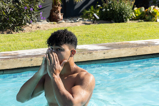 African American man 20s shirtless, standing chest-deep at backyard pool, raising hands to face