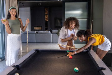 Obraz premium African American female friends leaning over black pool table lining shot with cue in living room