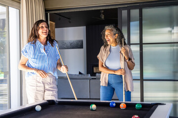 Obraz premium Diverse female friends chatting and playing on black-felt table in living room holding cues