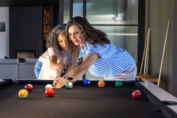 Obraz premium Diverse female friends leaning over black-felt pool table in rec room, pointing at billiard balls
