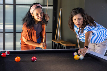 Naklejka premium Diverse female friends playing pool at black-felt table in rec room, holding cues and colored balls